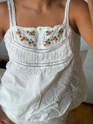 Linnetopp med spets - Säljer en så söt vit linnetopp med broderade blommor i flera färger på bröstet och spetsdetaljer. Toppen har smala axelband, knappar framtill och en luftig passform. 