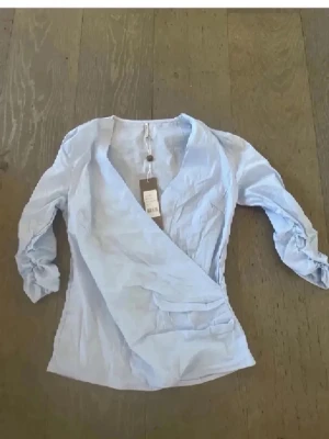 Ljusblå omlottblus med v-ringning - Snygg ljusblå omlottblus med djup v-ringning och långa ärmar. Blusen har en figurnära passform och är tillverkad i ett tunt, mjukt tyg som ger en elegant look. Perfekt att matcha med både jeans och kjol för en trendig stil. Passar även S