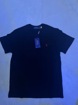 Svart t-shirt från Polo Ralph Lauren - Klassisk svart t-shirt från Polo Ralph Lauren med korta ärmar och rund hals. På bröstet finns den ikoniska röda logotypen broderad. Perfekt basplagg som passar till allt och är tillverkad i mjuk bomull för skön komfort.