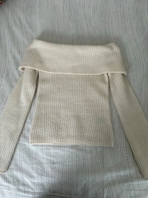 Off shoulder ribbad stickad tröja - Krämvit ribbstickad off shoulder-tröja med långa ärmar. Tröjan har en bred, vikt krage som ger en trendig look och är gjord i mjukt material som känns skönt mot huden. Perfekt för dig som vill ha en stilren och modern stickad topp. Endast använt en gång!