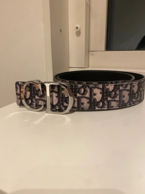 Svart och beige mönstrat bälte - Snyggt bälte med svart och beige mönster och en stor silvrig D-formad metallspänne. Bältet har ett grafiskt tryck längs hela bandet och känns riktigt trendigt. Perfekt accessoar för att lyfta vilken outfit som helst.