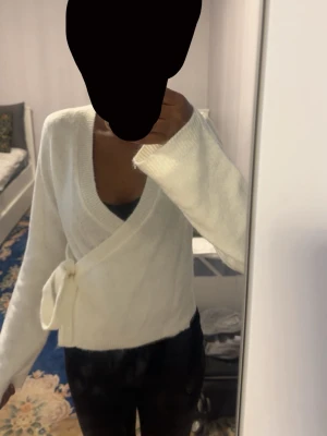 Vit omlottkofta med knytband - Mysig vit omlottkofta med knytband i midjan och långa ärmar. Koftan har en djup v-ringning och är tillverkad i ett mjukt, stickat material som känns skönt mot huden. Perfekt för lager på lager och enkel att styla till olika outfits.