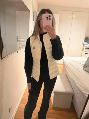 Vit dunväst med Moncler-logga - Snygg och lätt vadderad dunväst från Moncler i krispigt vit färg. Västen har hög krage, tryckknappar framtill och Monclers ikoniska logga på bröstet. Perfekt att slänga över en hoodie eller tröja för en trendig lager-på-lager-look. Storlek 14 passa mig som bruka ha S. Knappt använd 