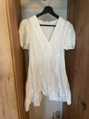 Vit klänning från Zara - Söt vit klänning från Zara med puffärmar och v-ringning. Klänningen har volangdetaljer och är tillverkad i ett lätt bomullstyg, perfekt för varma dagar. Enkel och stilren design som ger en fräsch look.