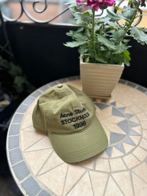 Olivgrön keps Acne Studios - Snygg olivgrön keps från Acne Studios med svart broderad text 'Acne Studios STOCKHOLM 1996' framtill. Klassisk böjd skärm och justerbar rem bak. Tillverkad i bomull för en bekväm och avslappnad look.