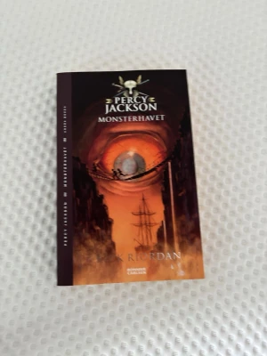 Percy Jackson: Monsterhavet bok - Spännande bok ur Percy Jackson-serien, 'Monsterhavet', skriven av Rick Riordan. Omslaget är svart och orange med ett öga och ett skepp i centrum. Boken är på svenska och ges ut av Bonnier Carlsen. Perfekt för dig som gillar äventyr och fantasy.