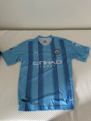 Manchester City matchtröja Haaland #9 - Snygg ljusblå Manchester City matchtröja från Puma med Haaland och nummer 9 på ryggen. Tröjan har klubbmärke på bröstet, Etihad Airways-tryck och klassiska ränder framtill. Perfekt för fotbollsfans och samlare.