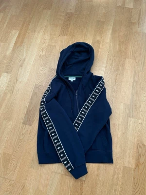 Mörkblå hoodie Lacoste 🐊 ”Äkta” - Snygg mörkblå hoodie med dragkedja och huva. Längs ärmarna går ett band med upprepat logotyp-tryck i vitt. Hoodien har fickor framtill och ribbade muddar. Perfekt för en chill och sportig look.