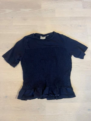 Mörkblå topp med volangkant - Snygg mörkblå topp med korta ärmar och smockad kropp som ger en tight passform. Toppen har en volangkant nedtill som ger en extra detalj. Perfekt att styla med jeans eller kjol för en trendig look.
