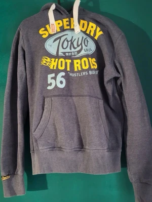 Grå Superdry hoodie Tokyo Hot Rods limited edition  - Snygg grå hoodie från Superdry med tryck i gult, vitt och blått framtill. Stora bokstäver, Tokyo Hot Rods-motiv och nummer 56. Huva med vita snören och känguruficka. Perfekt för en avslappnad streetstyle-look. Lite slitning på märket 