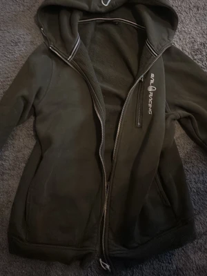 Mörkgrön hoodie från Sail Racing - Mörkgrön hoodie från Sail Racing med dragkedja framtill och broderad logga på bröstet. Hoodien har en stor huva och mjukt fleecefoder på insidan. Perfekt för dig som gillar en sportig och avslappnad stil.