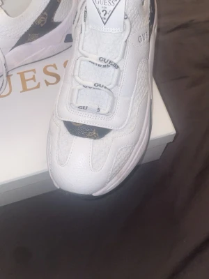Vita sneakers från Guess - Stilrena vita sneakers från Guess med svarta och guldiga logodetaljer på hälen och tungan. Skorna har en chunky sula, meshpaneler och vita snören med Guess-tryck. Perfekta för dig som gillar en sportig och trendig look.