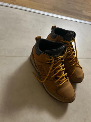 Bruna kängor från Helly Hansen - Säljer ett par bruna kängor från Helly Hansen med gula snören och svart vadderad kant. Kängorna har en robust sula och HH-logga på sidan. Tillverkade i slitstarkt material, perfekta för höst och vinter. Sköna att gå i och passar till många olika stilar.