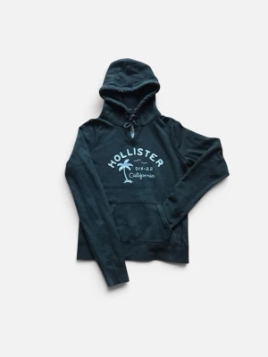 Mörkgrön Hollister hoodie med tryck - Snygg mörkgrön hoodie från Hollister med klassiskt tryck framtill och en palm. Hoodien har huva med dragsko och en stor magficka. Perfekt för chill dagar och enkel att matcha med jeans eller mjukisbyxor.