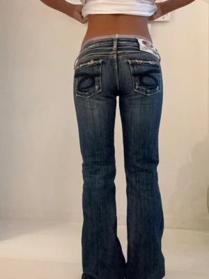 Blå bootcut jeans  - Säljer ett par blå bootcut jeans med snygga slitningar och broderade detaljer på bakfickorna. Jeansen har låg midja och klassisk femficksmodell. Perfekta för dig som gillar en avslappnad men ändå trendig look. W27 L32