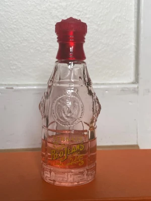 Versace Red Jeans Woman parfym - Versace Red Jeans Woman parfym i en genomskinlig glasflaska med detaljerad relief och röd plastkork. Flaskan har gul text och en lätt rosa ton på vätskan. Designen är retro och sticker ut med sin unika form och färgkombination.