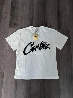 Vit corteiz tshirt - Snygg vit t-shirt med ett stort svart graffiti-inspirerat tryck på bröstet. Klassisk passform och rund halsringning. Tillverkad i mjuk bomull som känns skön mot huden. Perfekt för dig som gillar streetwear och vill sticka ut lite extra.