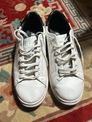 Vita sneakers med svart detalj - Stilrena vita sneakers med svart vadderad häl och vita snören. Klassisk låg modell med rund tå och diskreta sömmar. Ovandelen ser ut att vara i slätt konstläder och sulan är vit. Perfekta för en clean och enkel look.