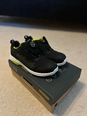 Svarta skyddskor från blåkläder med neon-detaljer - Snygga svarta sneakers från Blåkläder med vita sulor och neon-gula detaljer på insidan och sulan. Skorna har en sportig look med mönstrade sidor och praktisk snörning. Perfekta för dig som gillar streetstyle och vill sticka ut lite extra. Helt nya. Har även flera andra i olika storlekar, bara höt av dig