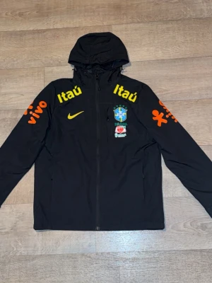 Svart Brasil Nike vindjacka med huva - Svart vindjacka med huva från Nike, dekorerad med Brasilien fotbollslandslagets logga och sponsortryck i gult och orange. Jackan har hel dragkedja framtill och två sidofickor. Materialet är tunt och vindtätt, perfekt för sportiga dagar.