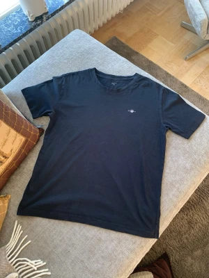 Mörkblå t-shirt från Gant - Snygg mörkblå t-shirt från Gant med liten logga på bröstet. Klassisk rund halsringning och korta ärmar. Tillverkad i mjuk bomull som känns skön mot huden. Perfekt till jeans eller shorts för en clean och enkel stil.
