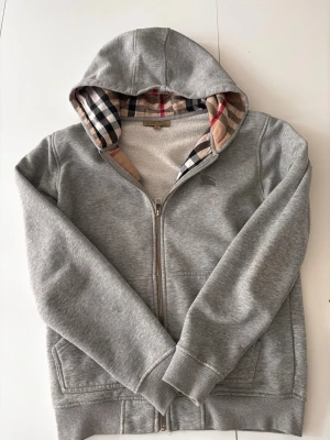 Grå hoodie med dragkedja från Burberry - Snygg grå hoodie från Burberry med dragkedja framtill och klassiskt rutigt foder i huvan. Tillverkad i mjukt material med ribbade muddar och fickor på sidorna. Perfekt för dig som vill ha en stilren och exklusiv look.
