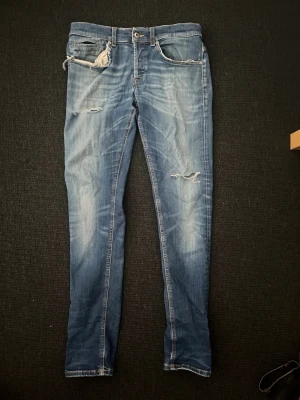 Blå slitna skinny jeans med dragkedja - Snygga blå skinny jeans med slitningar på låret och knät samt dragkedjedetalj vid fickan. Jeansen har klassisk femficksdesign, ljus tvätt och smal passform. Perfekta för en trendig och avslappnad look.