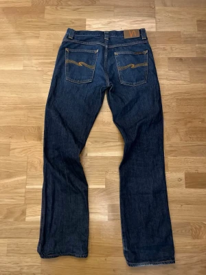 Mörkblå jeans från Nudie Jeans - Säljer ett par mörkblå jeans från Nudie Jeans med klassisk rak passform. Jeansen har kontrastsömmar och två bakfickor med Nudie Jeans karakteristiska broderi. Tillverkade i slitstark denim med läderpatch i midjan.