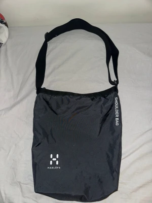 Haglöfs shoulder bag - Snygg svart axelväska från Haglöfs med justerbar rem och minimalistisk design. Väskan har tryckt logga framtill och texten 'SHOULDER BAG' på sidan. Tillverkad i slitstarkt syntetmaterial, perfekt för dig som vill ha en enkel och stilren väska.
