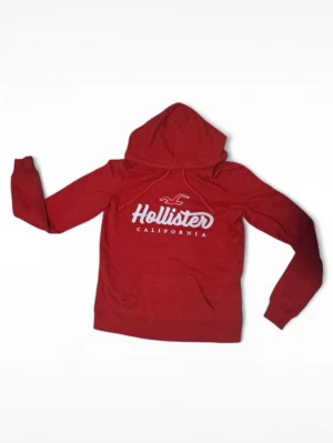Röd hoodie från Hollister - Snygg röd hoodie från Hollister med stor vit logga och texten 'California' på bröstet. Klassisk känguruficka framtill och huva. Perfekt för dig som gillar en avslappnad och sportig stil. Mjuk bomullsblandning som känns skön mot huden.