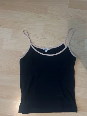 Mörkblått linne från Subdued - Säljer en svart linnetopp från Subdued med tunna axelband och beige kant runt halsringning och axelband. Enkel och stilren design som passar perfekt till sommaren. Toppen är ärmlös och har en slim passform.