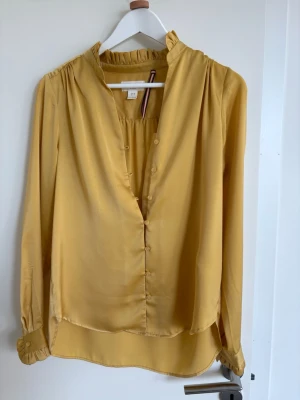 Gul blus från Holly & Whyte - Snygg gul blus från Holly & Whyte med volangkrage och knappar framtill. Blusen har lång ärm med knapp vid ärmslut och är tillverkad i ett glansigt, mjukt material som ger en lyxig känsla. Perfekt att styla till jeans eller kjol.