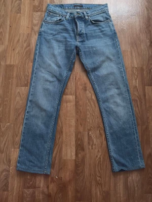 Nudie Jeans Grim Tim True Gold W30 L32 Slim Fit - Nudie Jeans “Grim Tim” i tvätten True Gold. Slim fit med lätt avsmalnande ben och normal midjehöjd – en av Nudies mest klassiska modeller. Storlek: W30 L32 Material: 99% bomull, 1% elastan Knappgylf, femficksmodell Skick: Mycket bra – inga hål, fläckar eller slitningar. Fin naturlig blekning i tvätten. Fler bilder finns – skriv gärna vid frågor!