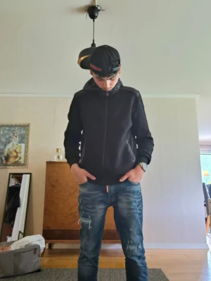 Svart zip-up cardigan med huva. Colmar cardigan köpte den på Zalando för 3500 kr så kom med ett rimligt pris  - Säljer en svart zip-up cardigan med huva och fickor framtill. Jackan har en enkel och clean design, perfekt för en avslappnad streetwear-look. Materialet ser ut att vara mjukt och bekvämt, och modellen är rak med långa ärmar.