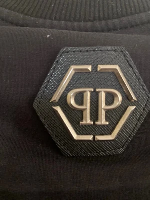 Svart hoodie med PP-logga - Snygg svart hoodie med en stor, sexkantig PP-logga i metall på ryggen. Hoodien har långa ärmar, ribbade muddar och är tillverkad i mjukt bomullstyg. Perfekt för dig som gillar stilrena och exklusiva detaljer.