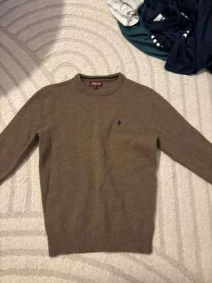 Beige stickad tröja från MCS - Snygg beige stickad tröja från MCS med rund halsringning och diskret mörkblå logga broderad på bröstet. Tröjan har ribbade muddar vid ärmslut och nederkant, perfekt för lager på lager och enkel att matcha med olika outfits.