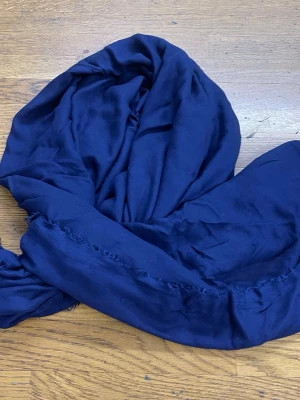 Mörkblå sjal med fransar - En stor, mjuk sjal i mörkblått med tunna fransar längs kanterna. Perfekt att svepa runt halsen för en stilren look. Materialet känns lätt och följsamt, vilket gör den enkel att styla på olika sätt.
