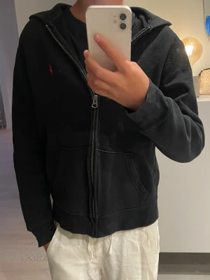 Svart hoodie från Ralph Lauren - Svart hoodie med dragkedja från Ralph Lauren. Klassisk design med huva, kängurufickor och röd broderad logga på bröstet. Tröjan är lite liten i storlek. Modellen är 158!
