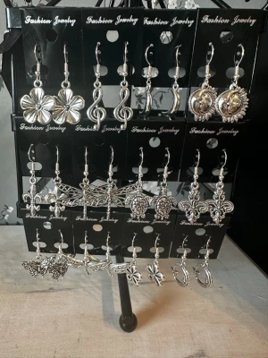 Set med 12 par silvriga örhängen - Säljer ett set med 12 par silvriga örhängen i olika former som blommor, noter, månar, fjärilar, drakflyg och ornament. Alla örhängen är i metall med snygga detaljer och hängande design. Perfekt för dig som vill variera din stil med coola accessoarer.
