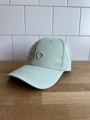 Ljusbeige/grön keps från Alo Yoga - Snygg ljusbeige keps från alo med broderad logga framtill. Klassisk böjd skärm och justerbar rem bak. Tillverkad i ett mjukt och tåligt tyg som passar perfekt till en avslappnad streetwear-look.