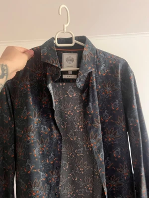 Mörkblå blommig skjorta från Stradivarius - Säljer en slim fit skjorta från Stradivarius i mörkblått med blommigt mönster i orange och grå toner. Skjortan har långa ärmar, klassisk krage och knappar framtill. Perfekt för dig som vill sticka ut med en unik stil.