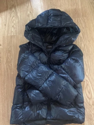 Marinblå pufferjacka med huva - Snygg marinblå pufferjacka med stor huva och quiltad design. Jackan har dragkedja framtill och breda kanaler som ger en riktigt puffig look. Perfekt för kalla dagar och riktigt varm. Materialet är glansigt och ger en modern vibe. Dam jacka men funkar absolut för män också