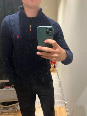 Mörkblå stickad tröja med dragkedja - Mörkblå stickad tröja från Ralph Lauren med halv dragkedja och klassiskt kabelstickat mönster. Tröjan har en röd broderad logga på bröstet och ribbade muddar vid ärmslut och nederkant. Perfekt för en stilren och avslappnad look.