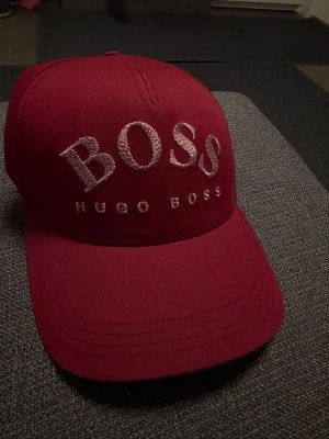 Vinröd keps från Hugo Boss - Snygg vinröd keps från Hugo Boss med stor broderad BOSS-logga framtill och mindre text under. Klassisk böjd skärm och justerbar baksida. Perfekt accessoar för en trendig och avslappnad stil.