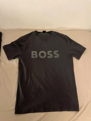 Svart BOSS t-shirt med tryck - Snygg svart t-shirt med stort BOSS-tryck på bröstet. Klassisk rund halsringning och korta ärmar. Perfekt för dig som gillar stilrena och enkla plagg med en tydlig logga. Tillverkad i mjukt material för skön känsla hela dagen.