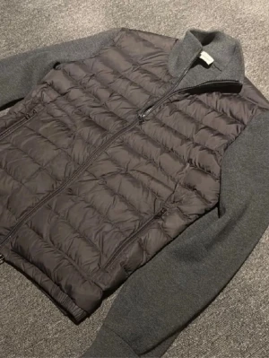 Moncler Cardigan  - Snygg grå pufferjacka med quiltad kropp och stickade ärmar samt hög krage. Jackan har dragkedja framtill och två sidofickor med dragkedja. Materialmix med vadderad kropp och mjuka, stickade detaljer på ärmar och krage. Perfekt för kalla vinterdagar.