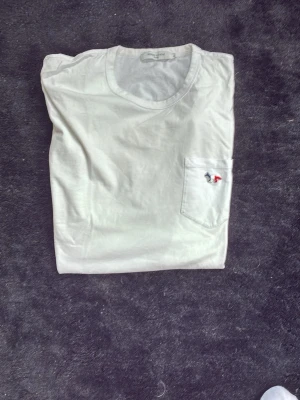 Vit t-shirt från Maison kitsune - Klassisk vit t-shirt i mjukt bomullstyg med rund halsringning och en bröstficka på vänster sida. På fickan finns en liten broderad detalj i rött och svart som ger en unik touch. Perfekt basplagg för alla tillfällen.