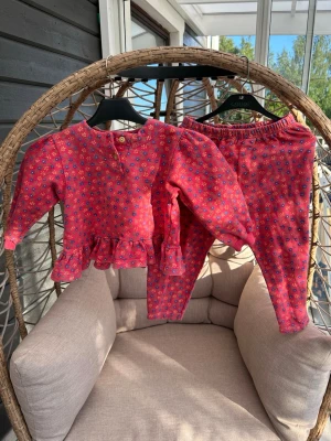 Loungewear-set strl. 98/104 - Sött loungewear-set i mjuk bomull från Robin. Setet består av en långärmad topp med volangkant och matchande byxor. Båda plaggen är rosa med små blommor i lila, gult och grönt.