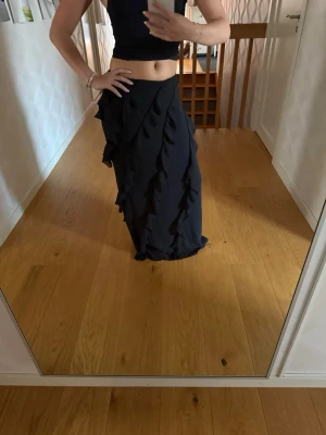 Svart långkjol med volangdetaljer - Säljer en svart långkjol med snygga volangdetaljer längs hela kjolen. Kjolen har en figurnära passform upptill och faller snyggt ner mot golvet. Perfekt för dig som vill ha en stilren men ändå unik look.
