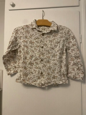Blommig skjorta från Newbie, strl 104 - Supersöt skjorta från Newbie i storlek 104 med blommigt mönster i beige, grönt och brunt. Skjortan har långa ärmar, klassisk krage och knappar framtill. Tillverkad i mjukt bomullstyg, perfekt för en söt och avslappnad look.